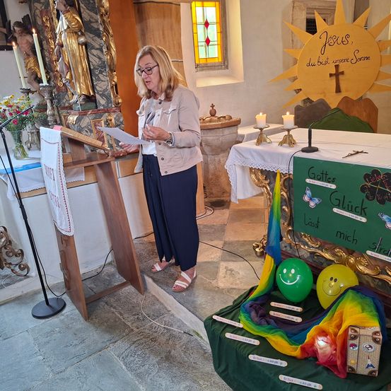 Eine Frau in einer Kirche spricht an einem Podest mit Kerzen, einer Statue und einem dekorierten Altar mit Luftballons und einem Regenbogentuch.
