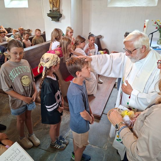 Ein Priester segnet Kinder in einer Kirche. Eine Frau hält einen Korb mit Blumen. Kinder sitzen auf Kirchenbänken.