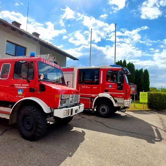 Zwei rote Feuerwehrfahrzeuge sind vor einem Gebäude mit klarem Himmel darüber geparkt. Eins hat das Wort 'Feuerwehr' auf der Seite.