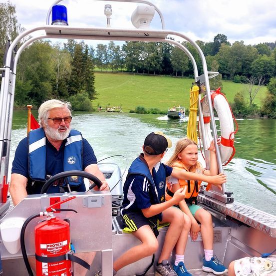 Ein älterer Mann fährt mit zwei Kindern, eines hält einen Feuerlöscher, auf einem ruhigen Fluss mit grünen Feldern und Bäumen im Hintergrund.