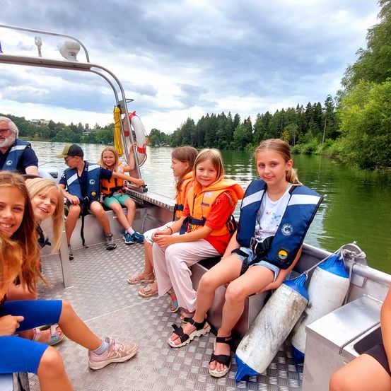 Mehrere Kinder und ein Erwachsener in Rettungswesten sitzen auf einem Boot auf einem See mit grünem Wasser und Bäumen am Ufer.