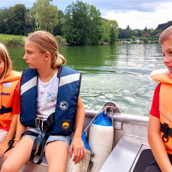 Drei Kinder in Schwimmwesten sitzen auf einem Boot auf einem Fluss. Bäume und Häuser sind im Hintergrund sichtbar.