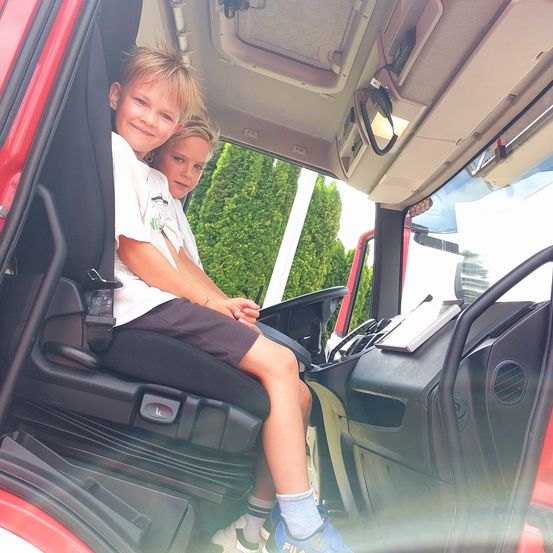 Zwei junge Jungen sitzen in einem roten Fahrzeug, möglicherweise einem Feuerwehrauto, mit grünem Baumhintergrund. Der Junge vorne trägt ein weißes Hemd und blaue Schuhe, während der Junge dahinter ein weißes Hemd und weiße Schuhe trägt.