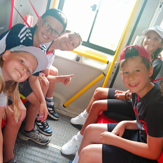 Eine Gruppe von Kindern sitzt lächelnd in einem Fahrzeug. Drei Jungen tragen Hüte und Turnschuhe, und ein Mädchen trägt eine weiße Kappe. Der Businnenraum hat gelbe Handläufe und ein Fenster.