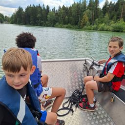 Drei Jungen auf einem Boot mit Schwimmwesten, lächeln in die Kamera. Hinter ihnen ist ein See mit Bäumen.