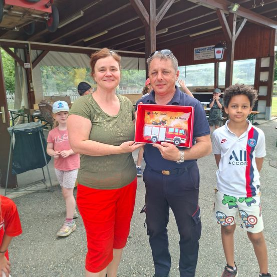 Ein Mann und eine Frau stehen mit einem Kind und halten eine Geschenkbox mit einem Feuerwehrauto-Bild. Hinter ihnen schaut ein Junge in einem Paris Saint-Germain-Trikot zu. Im Hintergrund ein Gebäude mit einem Schild und einer Person.