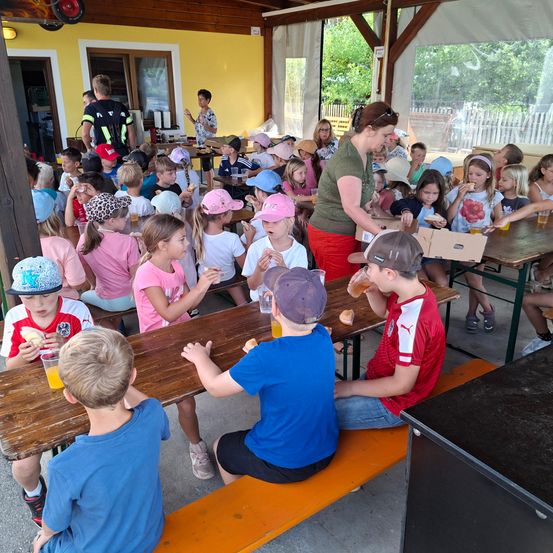 Eine Gruppe von Kindern sitzt an einem Tisch und isst und trinkt. Eine Frau serviert ihnen Essen.