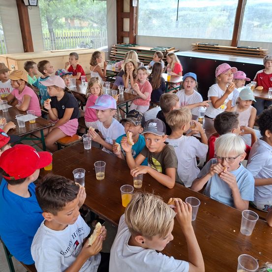 Eine Gruppe von Kindern in einem Essbereich isst Sandwiches, einige trinken aus Plastikbechern, alle tragen Mützen.