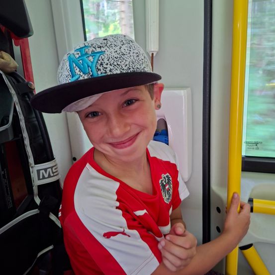 Ein junger Junge lächelt, während er einen gelben Handgriff an einem Fahrzeug hält. Er trägt ein rotes und weißes Shirt mit einem Logo und eine Mütze mit NY darauf.