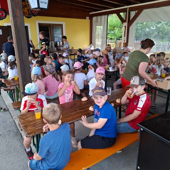 Kinder sitzen an einem Picknicktisch und essen. Eine Frau in Grün steht hinter dem Tisch. Viele Kinder sitzen an Tischen in einem Pavillon.