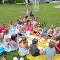 Eine Gruppe von Kindern sitzt auf einer Decke im Park, isst Snacks und macht ein Picknick. Ein Fußballtor ist im Hintergrund.