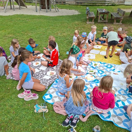 Eine Gruppe von Kindern sitzt auf einer Decke in einem Park und isst. Einige tragen Hüte und Turnschuhe. Hinter ihnen steht eine Holzbank und ein Tisch. Ein Rutsche und ein Zaun befinden sich auf der linken Seite.