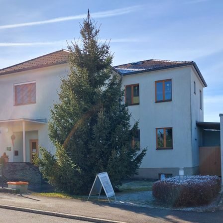 Ein modernes Haus mit braunem Dach und Solarpanels. Ein großer Baum und ein Schild stehen davor.