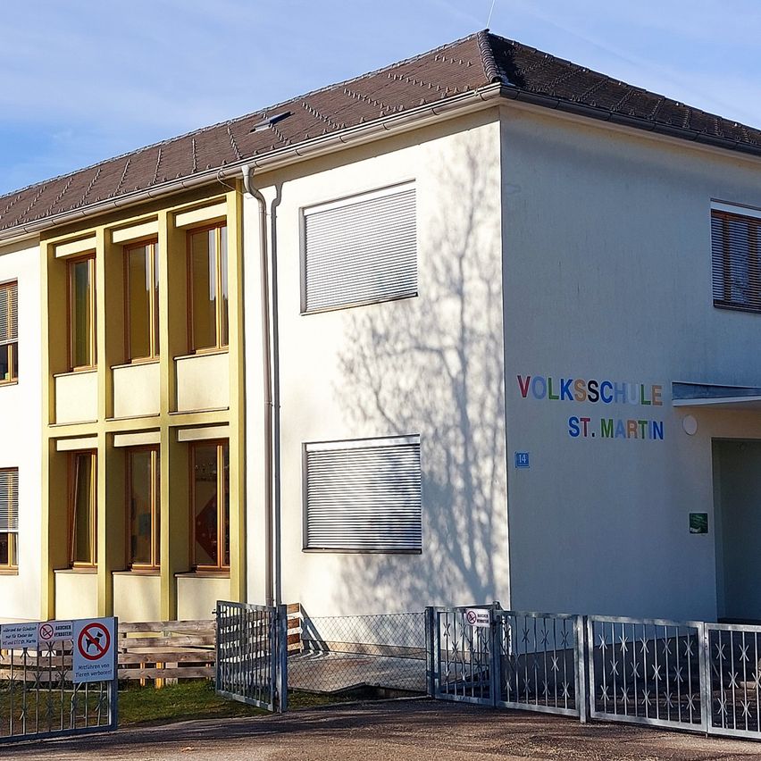 Ein weißes Gebäude mit gelben Akzenten namens Volksschule St. Martin hat ein braunes Dach, mehrere Fenster und zwei Tore vorn. Es gibt zwei Schilder vor dem Gebäude.