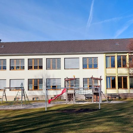 Ein großes Gebäude mit vielen Fenstern und einem Spielplatz davor. Der Spielplatz hat eine rote Rutsche und eine Kletterstruktur. Das Gebäude hat ein graues Dach.