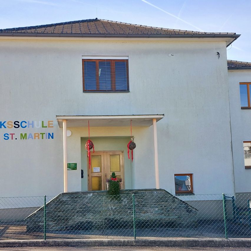Die Vorderseite eines Schulgebäudes mit der Aufschrift 'KSSCHULE ST. MARTIN' mit einer Holztür und zwei Fenstern. Ein grüner Zaun umgibt den Eingang, und eine kleine Weihnachtsdekoration steht davor.