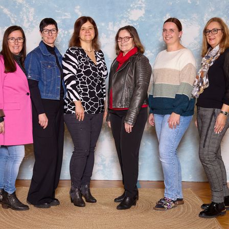 Eine Gruppe von sechs Frauen in verschiedenen Outfits und Accessoires posiert für ein Foto vor einem blauen Hintergrund mit Wolken.