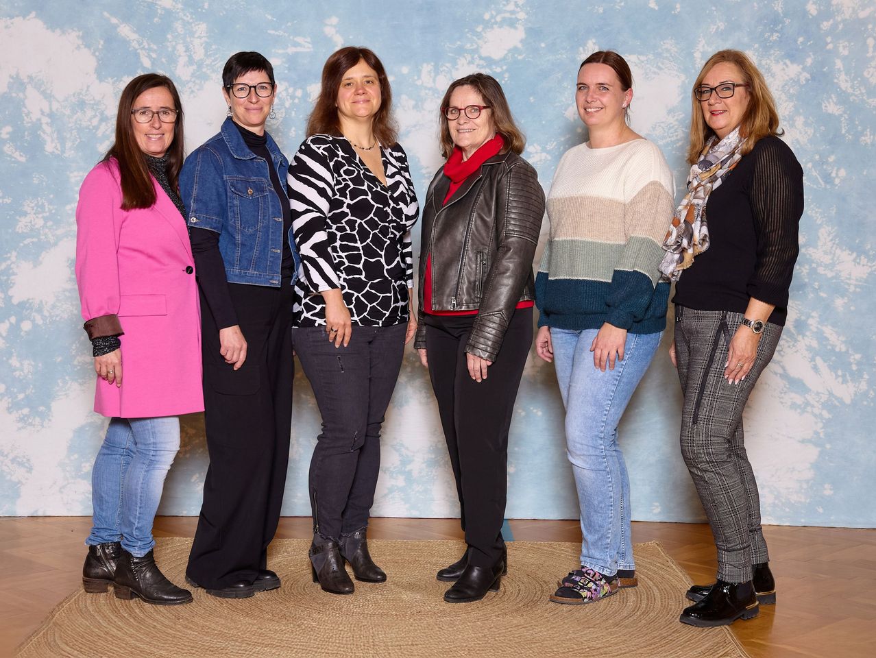 Eine Gruppe von sechs Frauen in verschiedenen Outfits und Accessoires posiert für ein Foto vor einem blauen Hintergrund mit Wolken.