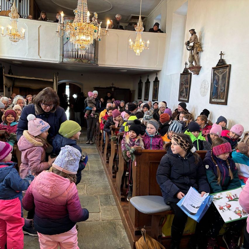 In einer Kirche sitzen Kinder auf Kirchenbänken, begleitet von Erwachsenen. Die Kirche hat Kronleuchter, Gemälde an den Wänden und eine Empore mit Menschen.