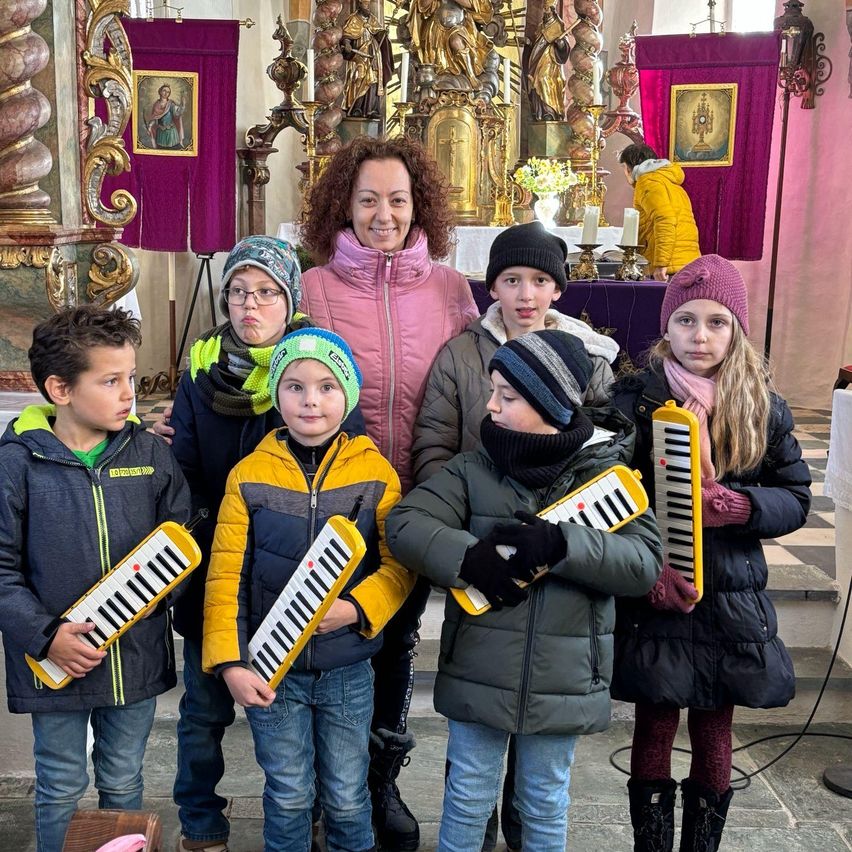 Eine Frau und mehrere Kinder stehen in einer Kirche und halten Spielzeug-Keyboards. Sie sind alle warm in Winterkleidung gekleidet. Hinter ihnen ist ein Altar mit Kerzen und religiösen Symbolen geschmückt.