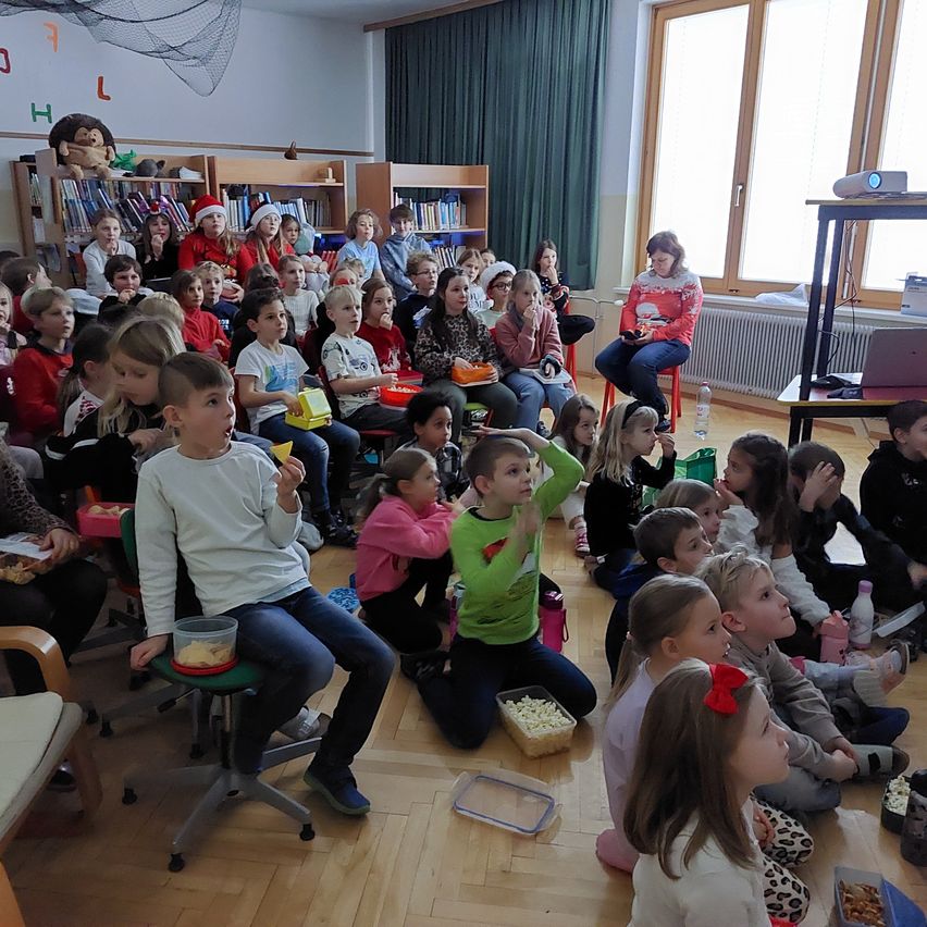 Ein Klassenzimmer voller Kinder, die einen Film sehen, einige auf Stühlen, andere auf dem Boden, und genießen Snacks. Ein Beamer steht auf einem Ständer, und eine Lehrerin sitzt in der Nähe.