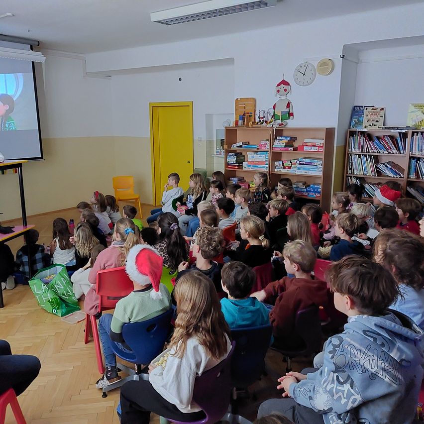 Kinder versammeln sich in einem Klassenzimmer und schauen sich einen Film auf einer Projektionsleinwand an. Einige tragen Weihnachtsmützen. Bücherregale und eine Uhr sind an der Wand. Stühle und ein Tisch stehen im Raum.