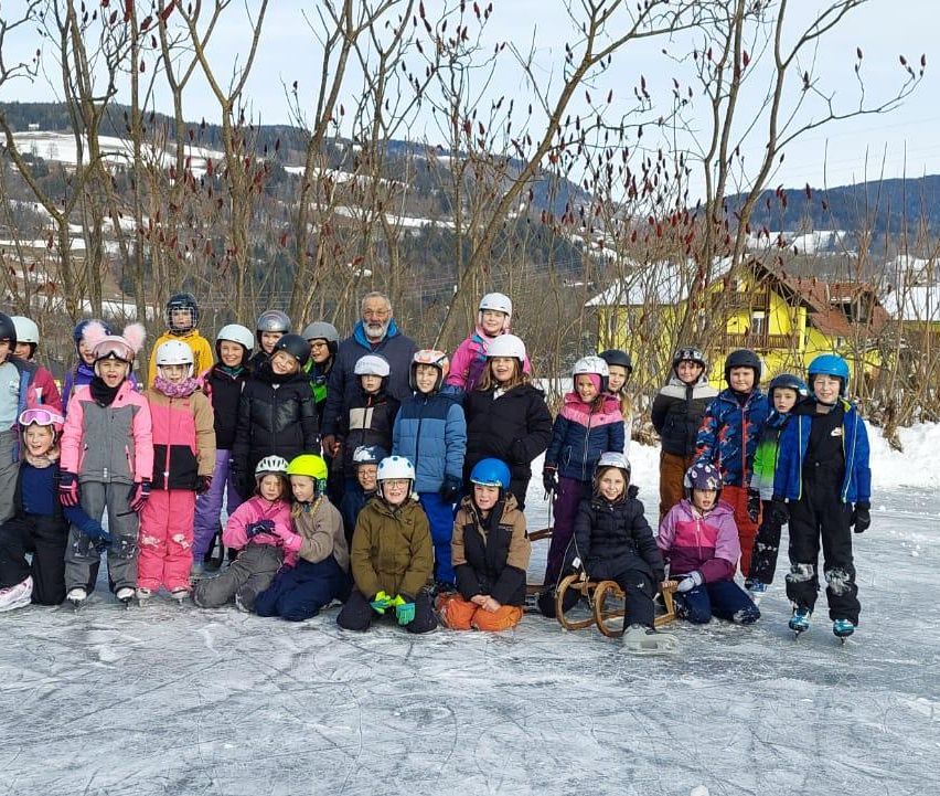 Eine Gruppe von Kindern und Erwachsenen steht und sitzt auf einer Eisbahn mit Winterkleidung und Helmen. Dahinter stehen kahle Bäume und ein gelbes Haus mit schneebedecktem Dach.