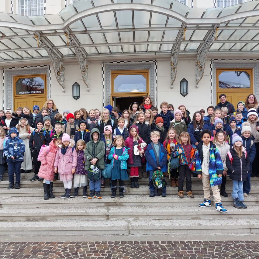 Eine große Gruppe von Kindern und Erwachsenen steht auf den Stufen eines historischen Gebäudes, einige tragen Mützen und Wintermäntel. Das Gebäude hat Glasfenster und kunstvolle architektonische Details.