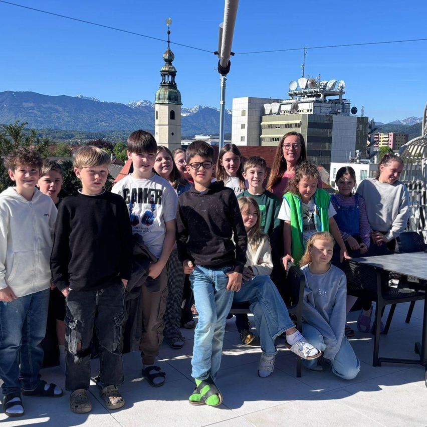 Eine Gruppe von Kindern und Erwachsenen steht lächelnd für ein Foto auf einer Dachterrasse. Dahinter befinden sich Gebäude und Berge in der Ferne.