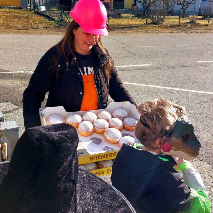 Eine Frau in einem rosa Schutzhelm hält eine Schachtel Donuts für ein Kind mit einer Dinosauriermaske.
