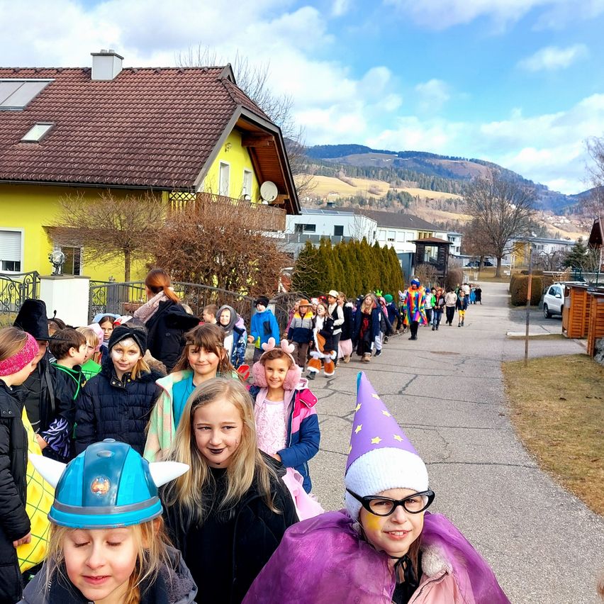 Kinder in Kostümen, darunter Hexen und Ritter, gehen in einer Reihe die Straße eines sonnigen Dorfes hinunter, mit Bergen im Hintergrund.