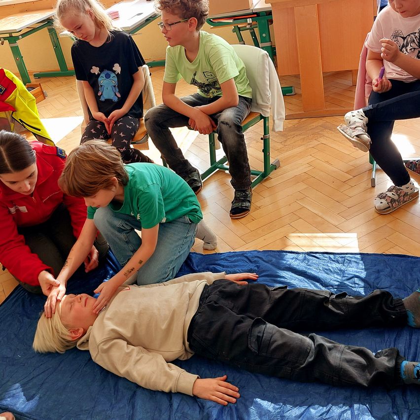 In einem Klassenzimmer üben mehrere Kinder Erste Hilfe an einem Übungsmodell. Einige sitzen auf Stühlen, während andere neben dem Modell kauern. Der Raum ist durch Sonnenlicht beleuchtet.