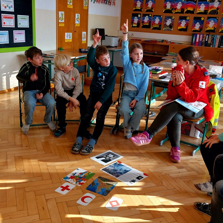 Kinder sitzen im Kreis auf dem Boden, einige mit erhobenen Händen. Eine Lehrerin sitzt neben ihnen. Papiere mit verschiedenen Symbolen und einer Globus liegen auf dem Boden.