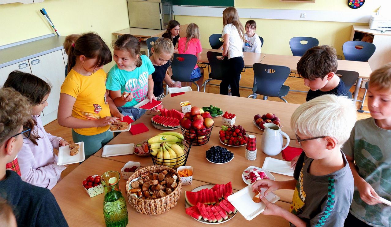 Eine Gruppe von Kindern ist um einen Tisch versammelt, auf dem verschiedene Lebensmittel stehen. Es gibt Wassermelonen, Bananen, Erdbeeren und andere Früchte auf dem Tisch. Einige Kinder essen, während andere stehen und das Essen betrachten.