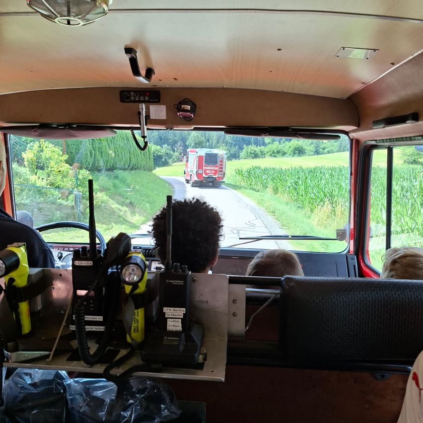 Ein Feuerwehrwagen fährt auf einer Straße, umgeben von Gras. In einem Fahrzeug sitzt eine Person mit Funkgerät und anderer Ausrüstung.