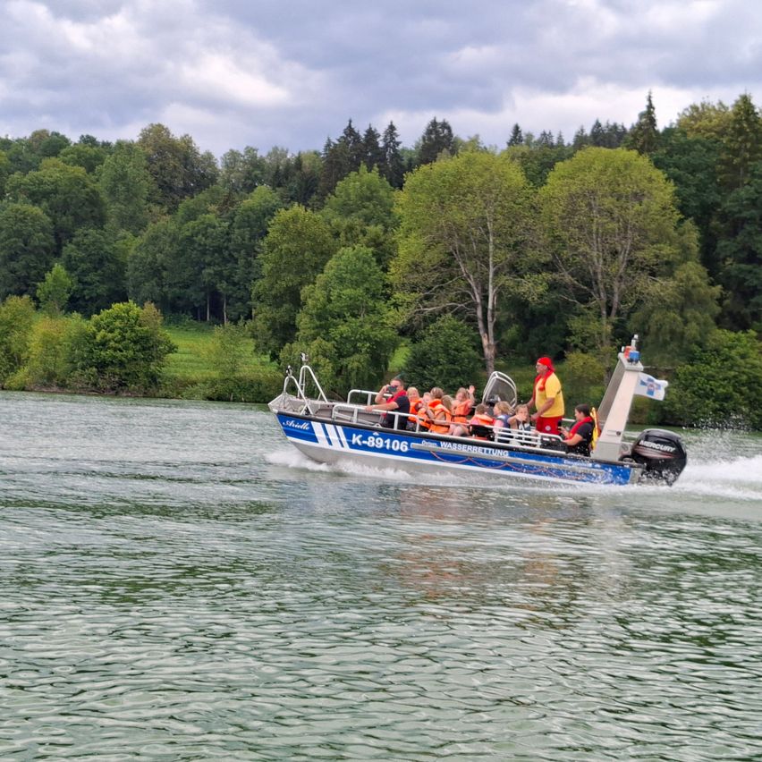 Ein Boot mit der Nummer K-89106 fährt mit Passagieren in Schwimmwesten auf einem Fluss. Bäume säumen den Fluss.