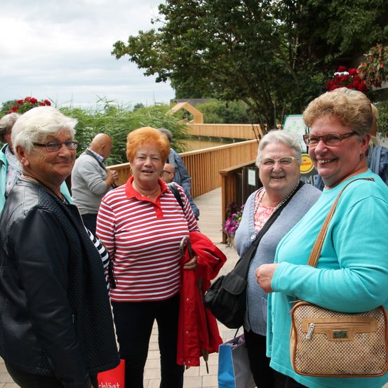 Vier Frauen stehen lächelnd auf einem Holzweg, mit einer Brücke und Grünpflanzen im Hintergrund. Eine Frau hält eine rote Jacke.