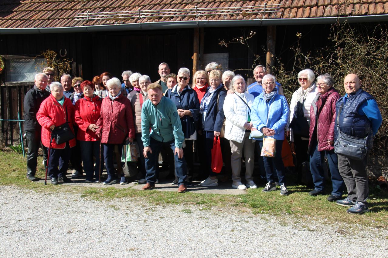 Eine Gruppe von Menschen steht vor einem Gebäude, manche halten Taschen, andere tragen Sonnenbrillen. Sie sind warm gekleidet, was auf kaltes Wetter hinweist.