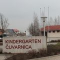 Ein Kindergartengebäude mit einem Schild, das Kindergarten Cuvamica steht, umgeben von kahlen Bäumen, Gras und geparkten Autos.