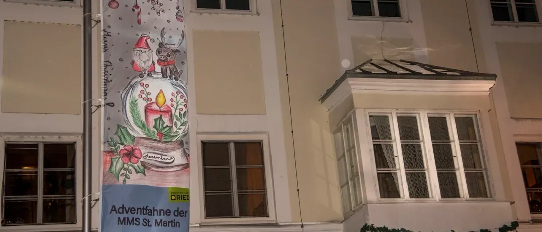 Eine Fahne mit der Nummer 15 und Weihnachtszeichnungen ist an einem beigefarbenen Gebäude aufgehängt. Die Fahne zeigt den Weihnachtsmann, ein Rentier und eine Kerze. Das Gebäude hat zwei Stockwerke, mit Fenstern auf jeder Etage. Das untere Stockwerk hat Weihnachtslichter auf der Brüstung.