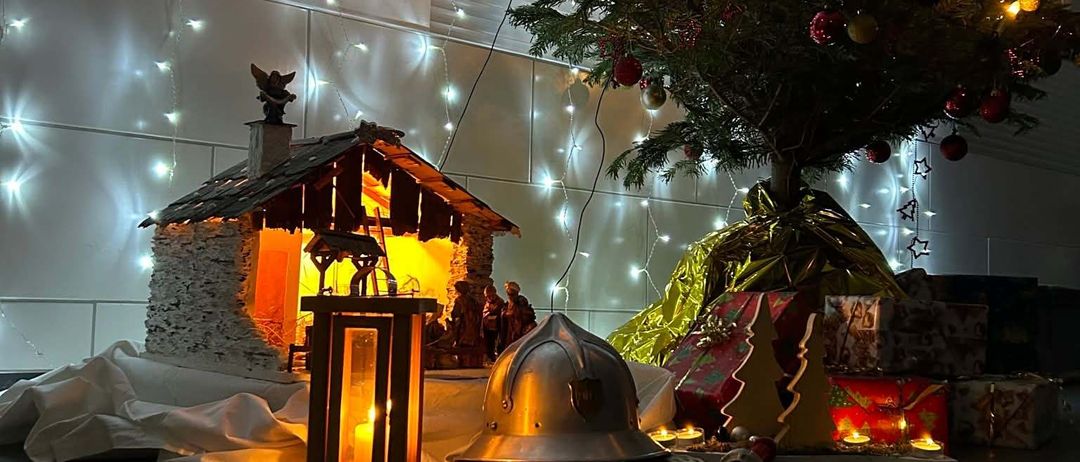 Ein Weihnachtsbaum mit bunten Lichtern und Ornamenten steht neben einer Krippe. Ein Feuerwehrhelm und eine Laterne sind in der Nähe, mit Geschenken und verpackten Präsenten um den Baum. Weiße Lichter hängen von der Decke.
