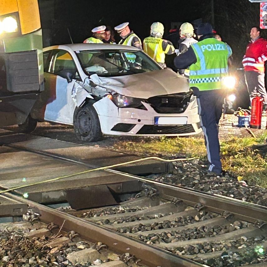 A white car with significant front damage is parked on a railway track. Emergency services, including police, are attending to the scene. A train is visible on the left side.