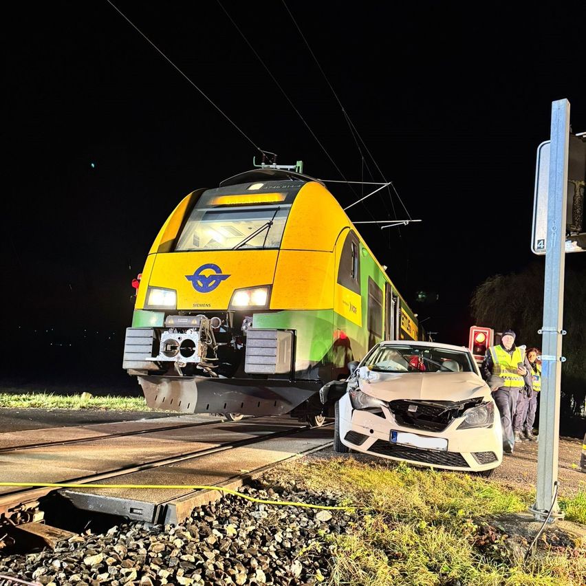A yellow train collided with a white car at night on a railway track. People in yellow vests stand nearby.