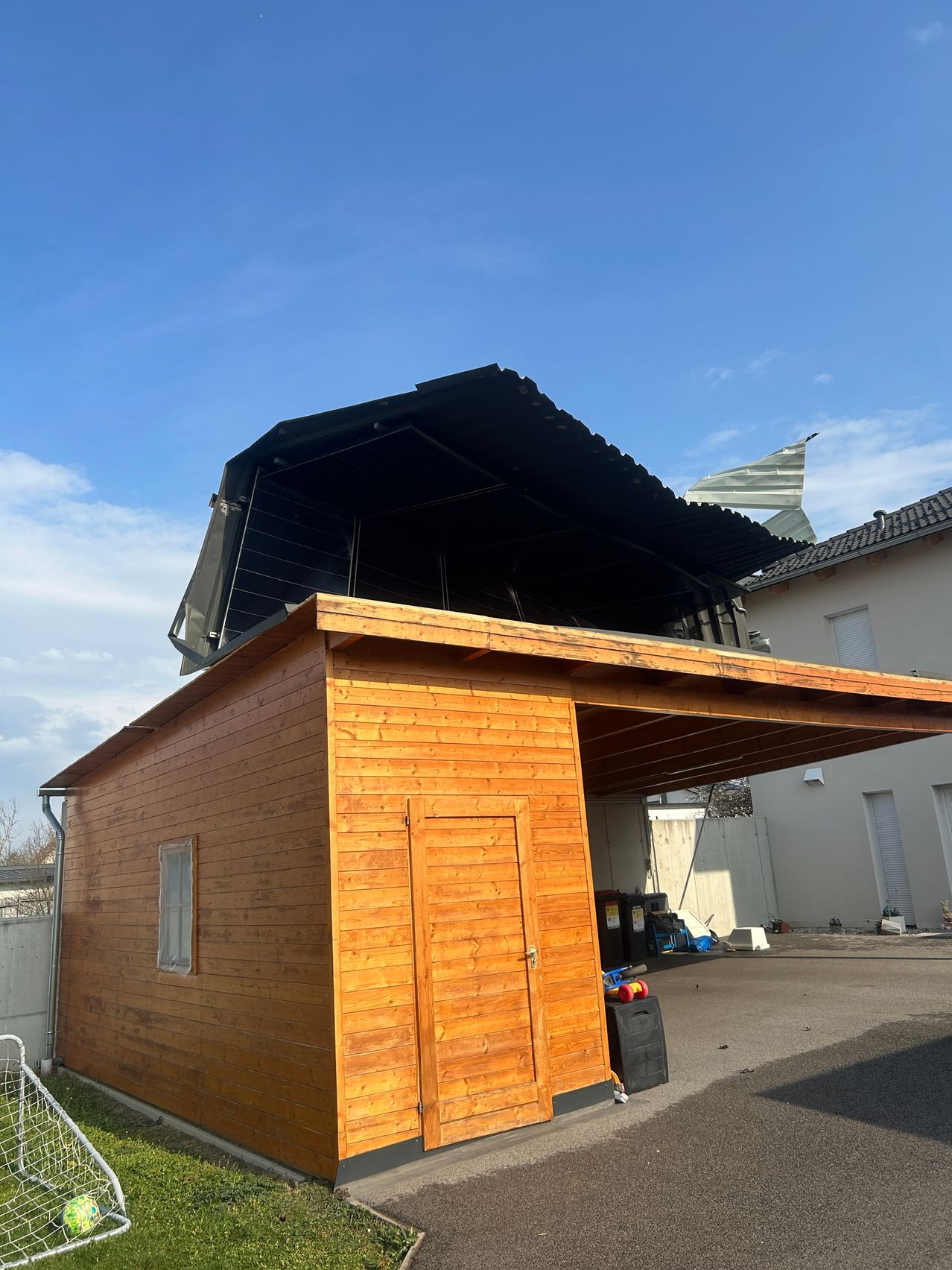 Ein hölzernes Schuppen mit Carport und schwarzem Vordach. Es ist ein sonniger Tag mit klarem blauem Himmel.