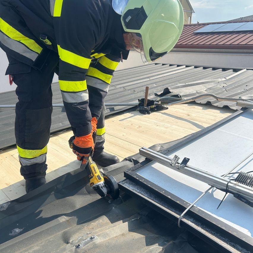 Ein Feuerwehrmann in einem grünen Helm und einer reflektierenden Schutzkleidung arbeitet auf einem Dach mit Solarmodulen. Er benutzt ein Elektrowerkzeug, um Anpassungen vorzunehmen.
