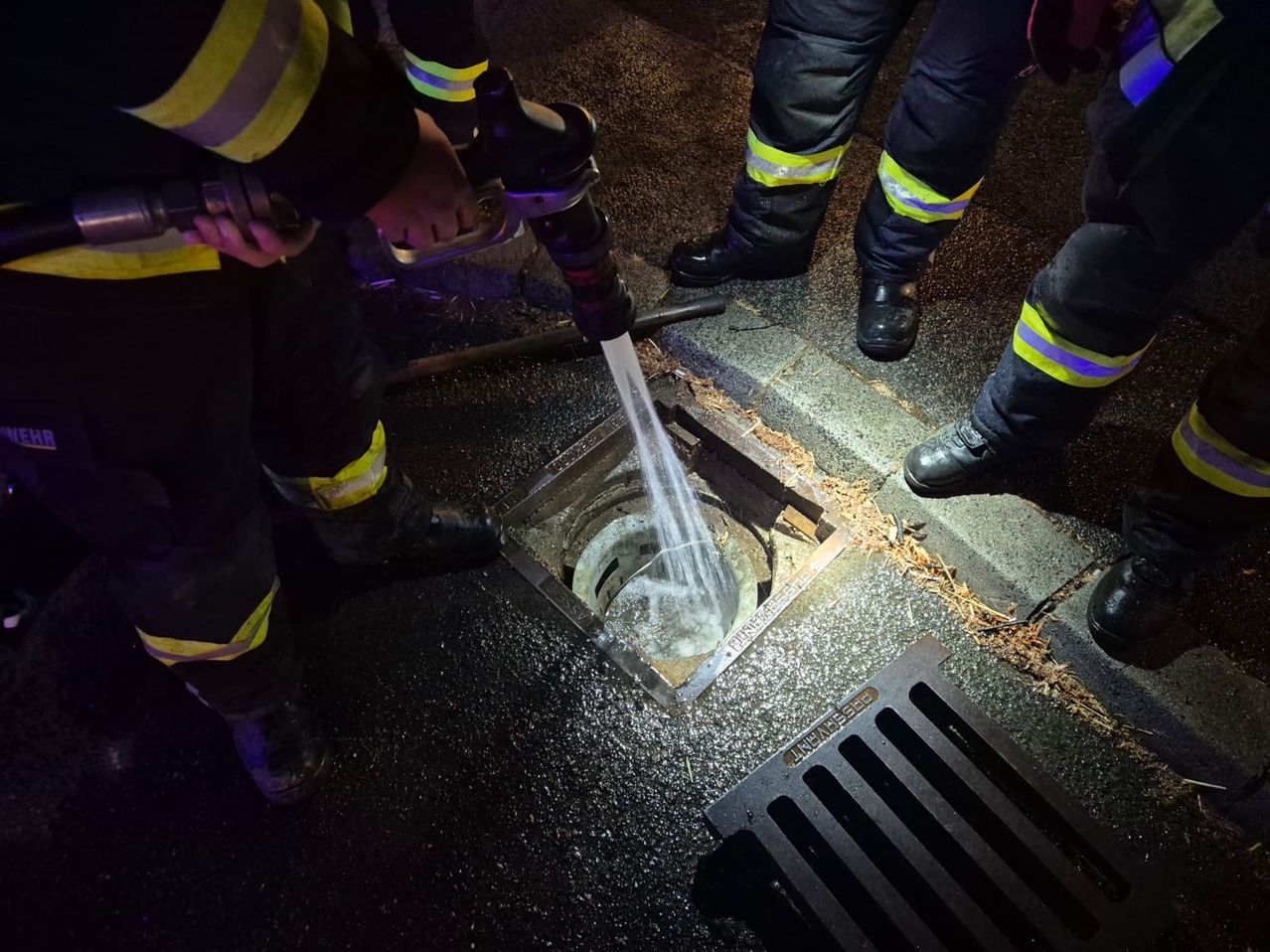 Firefighters are using a hose to clean a sewer grate on a wet street at night.
