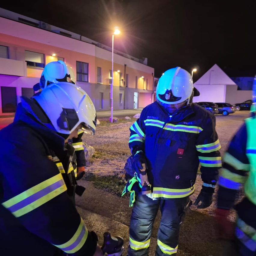 Firefighters in protective gear stand outside a building at night, one holding a tool.