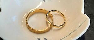 Eine weiße Keramik-Herzschale mit zwei goldenen Ringen darin, auf einer Holzoberfläche mit einer Lupe und einem Meißel.