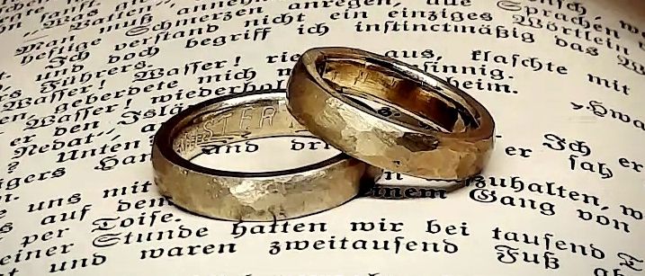 Zwei goldene Eheringe liegen auf einem offenen Buch mit deutschem Text, auf einer weißen Feder platziert. Die Ringe überlappen sich leicht.
