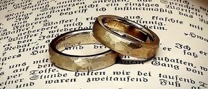 Zwei goldene Eheringe liegen auf einem offenen Buch mit deutschem Text, auf einer weißen Feder platziert. Die Ringe überlappen sich leicht.